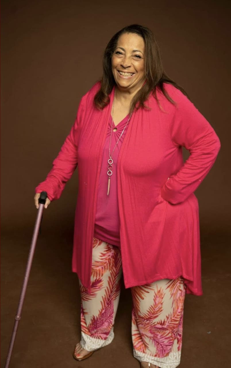 A smiling woman in a bright pink cardigan, resting on a cane, against a brown backdrop
