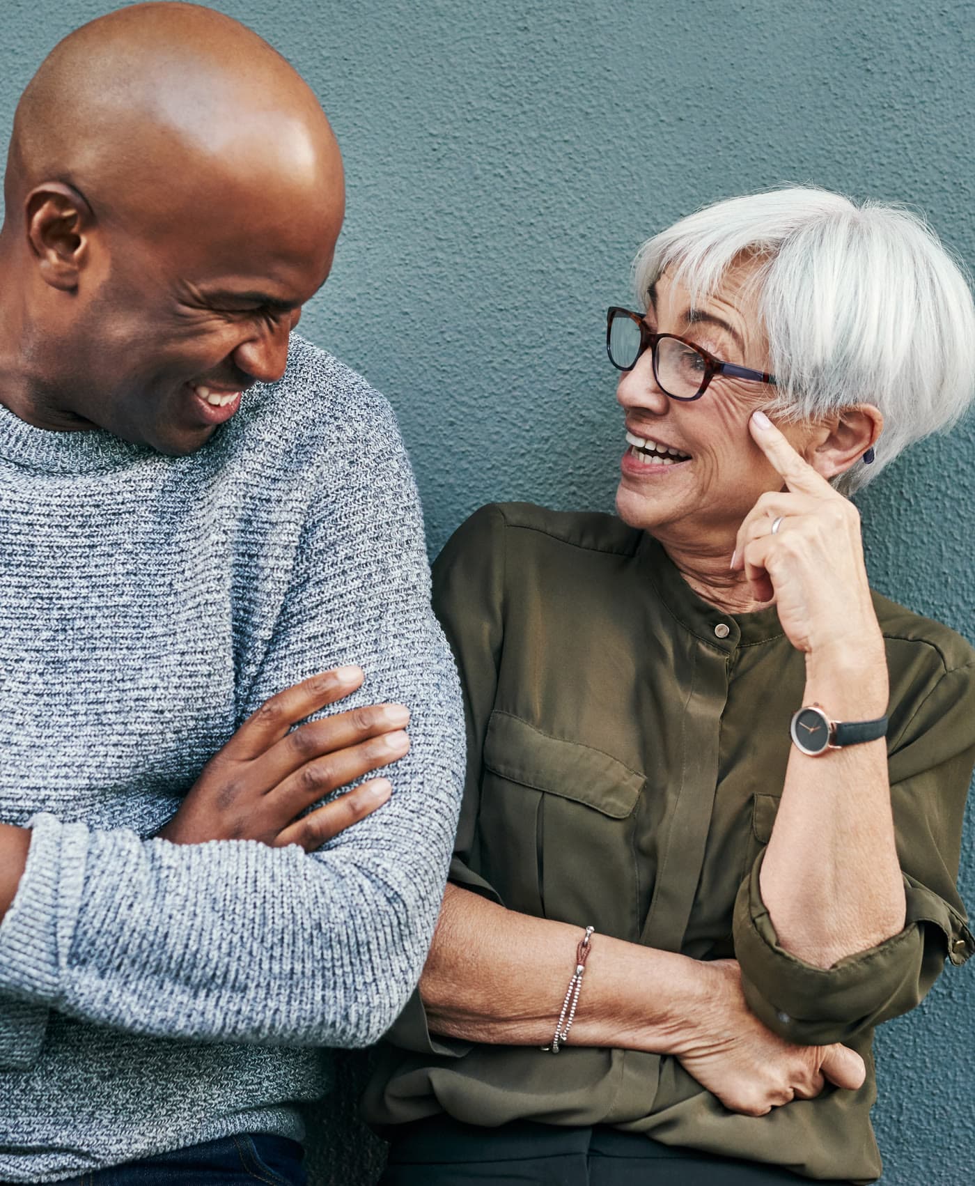 Two people laughing together against a blue-grey wall, one with short white hair