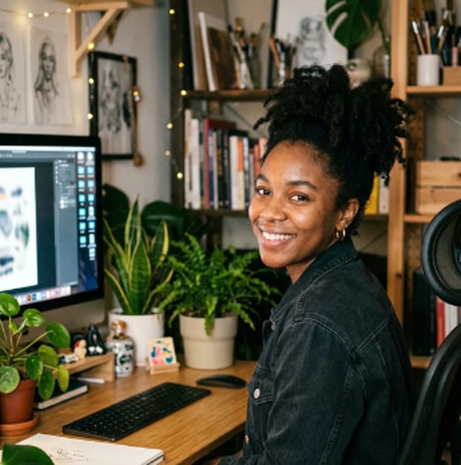 A smiling person seated at a desk with plants, a keyboard and a monitor