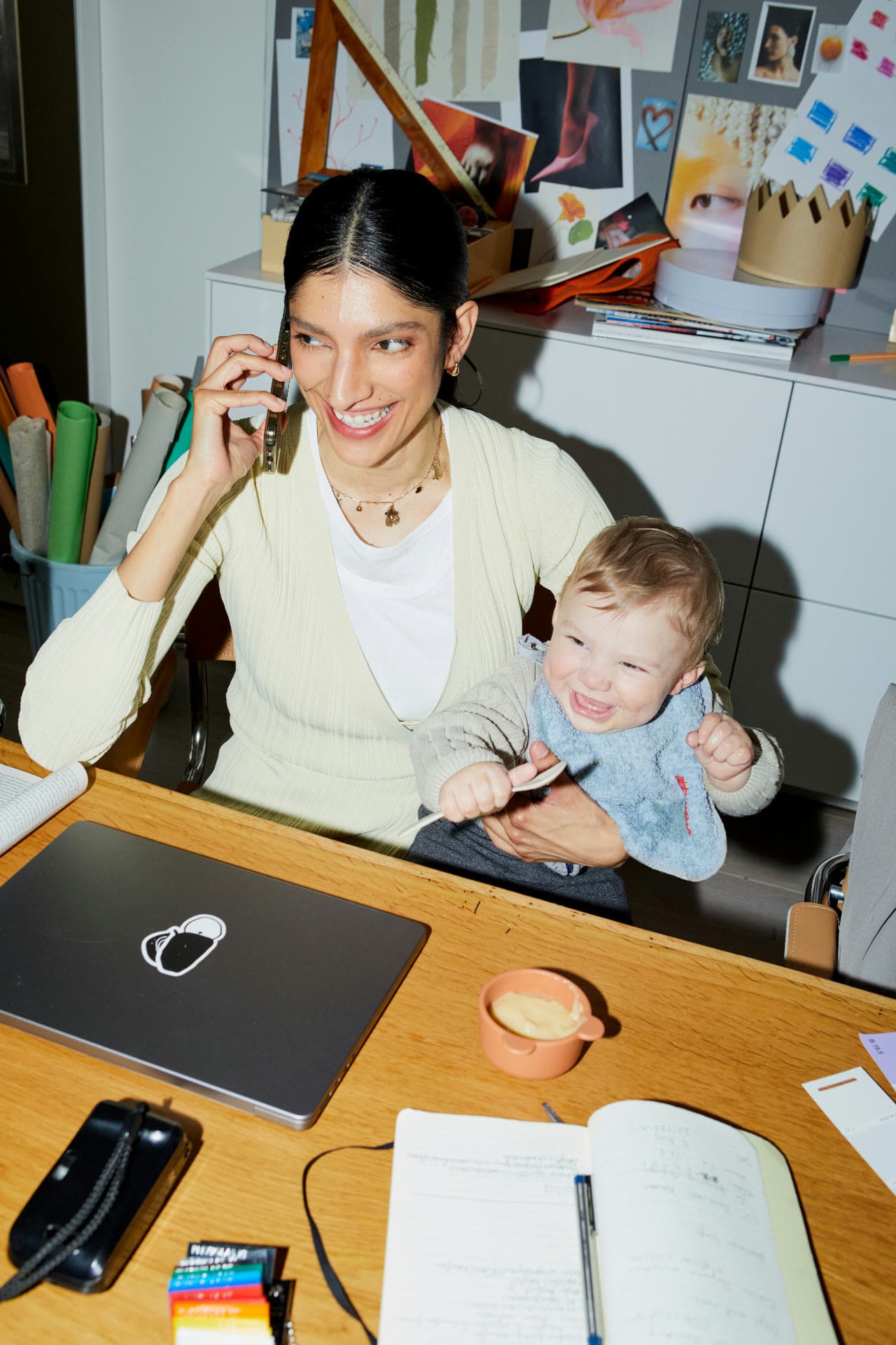 A smiling woman on a phone call at her desk with a laughing toddler on her lap